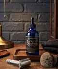 Blue bottle of 'Royal Legacy' body oil on a wooden tray with a razor and brush against a brick wall background.