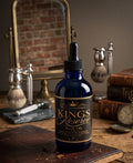 Blue bottle of 'Kings Reserve' body oil on a wooden surface with vintage shaving tools and books in the background.