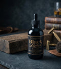 Bottle of 'Black Label' body oil on a dark surface with vintage books and tools in the background.