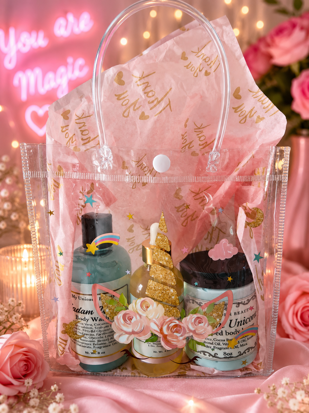 A clear plastic gift bag filled with pink tissue paper and assorted bath/body treats, including a blue bottle, a gold unicorn-shaped candle, a pink flower decal, and a jar, styled with pink roses and fairy lights.