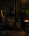 Two black labeled bottles of 'My Unicorn Beauty' Black Label Body Wash and Body Oil on a dark surface with decorative elements.