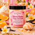 A jar of My Unicorn Beauty Unicorn Rainbow Orange Cream Vanilla Whipped Sugar Scrub is shown in a styled setting with orange slices, white flowers, vanilla beans, and whipped cream, with sugar scattered around.