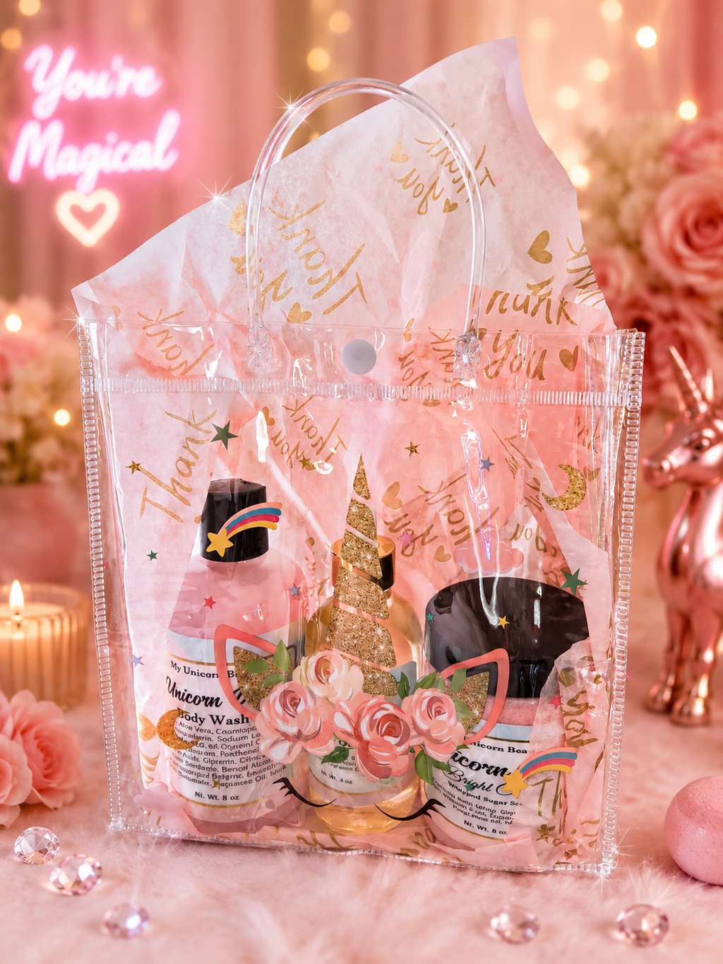 A pink and gold themed gift set in a clear plastic bag with a handle, containing a body wash, a cream product, and decorative paper, styled with roses, candles, and fairy lights.