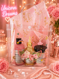 A pink unicorn-themed gift set in a clear plastic tote bag with gold foil details, surrounded by pink roses, pearls, and candles. The set includes a pink body wash, a dark chocolate brown body scrub, and a small gold unicorn figurine, with pink tissue paper printed with 'Unicorn Dreams'.