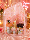 A pink unicorn-themed gift set in a clear plastic tote bag with gold foil accents, displayed on pink satin fabric with decorative flowers, candles, and a gold unicorn figurine in the background.