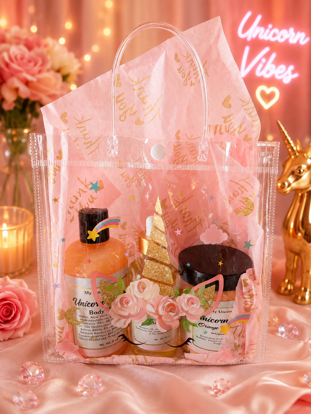 A pink unicorn-themed gift set in a clear plastic tote bag with gold foil accents, displayed on pink satin fabric with decorative flowers, candles, and a gold unicorn figurine in the background.